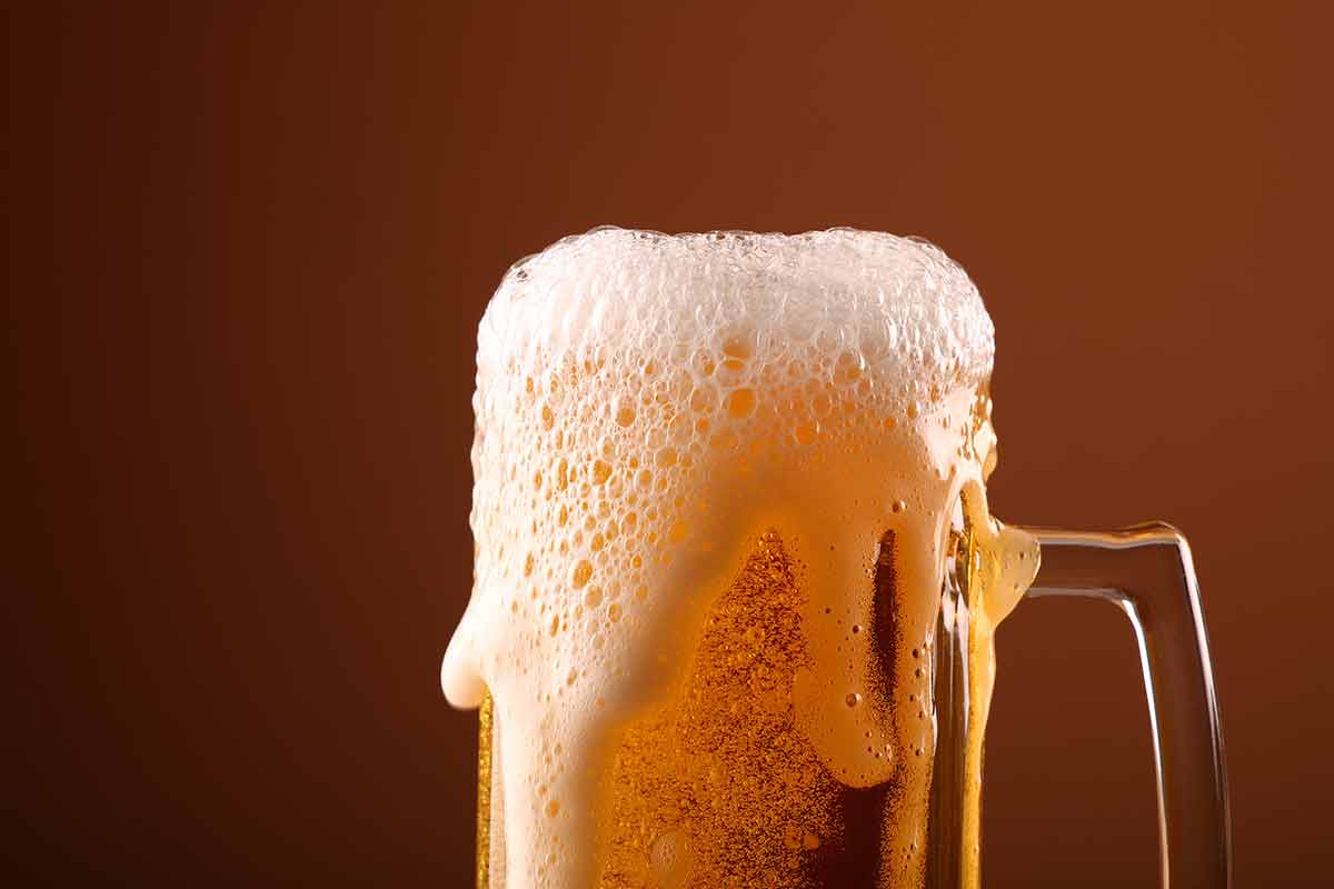 American beer frothy close up pouring lager beer with white froth and bubbles in glass mug over dark brown background with copy space, low angle side view