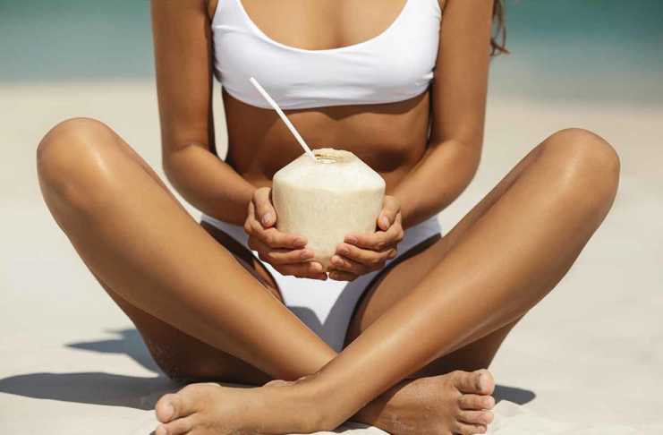 20 Things To Do In Malibu That Go Beyond Celebrity Spotting Malibu tanned woman in a white sports bikini sitting crossed-legged on the sand drinking juice from a coconut