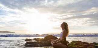 Woman sitting cross-legged on rocks by the beach meditating
