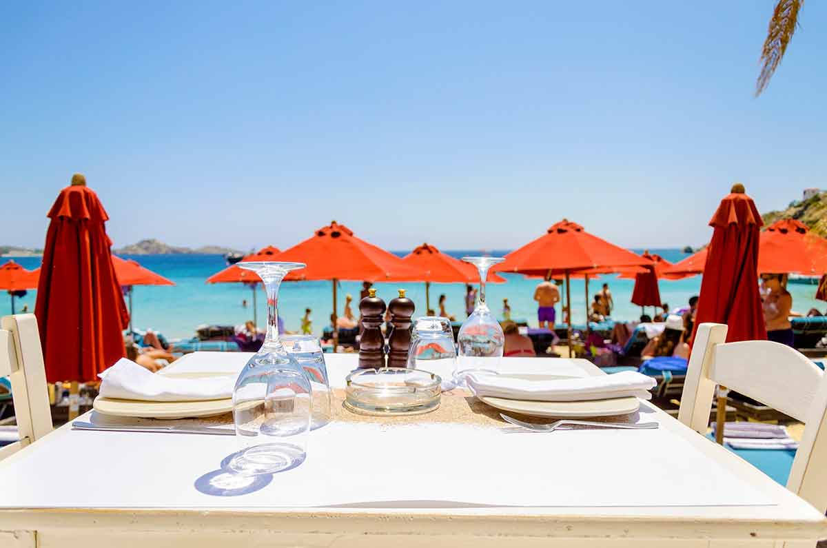 best beaches in mykonos for families A table set with plates, glasses and napkins on a beach bar restaurant and a view of the sun umbrellas and the blue sea