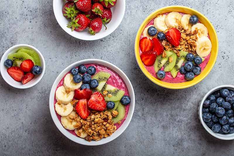 best cafes in amsterdam Summer acai smoothie bowls with strawberries, banana, blueberries, kiwi fruit and granola on gray concrete background.