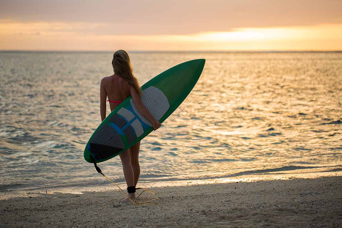 best swimming beaches in hawaii surfer girl looking at ocean holding a surfboard at sunset