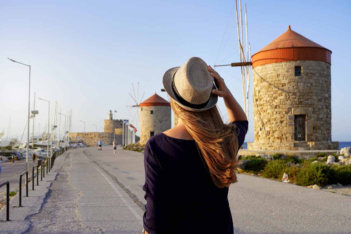 best time to visit greece Back view of young traveler woman walking along promenade with old windmills of Rhodes, Europe. Sunset.