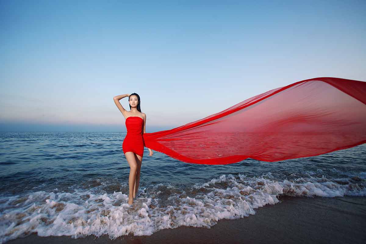 china beaches model wearing a red dress and flowing scarf standing on the beach among the waves. 