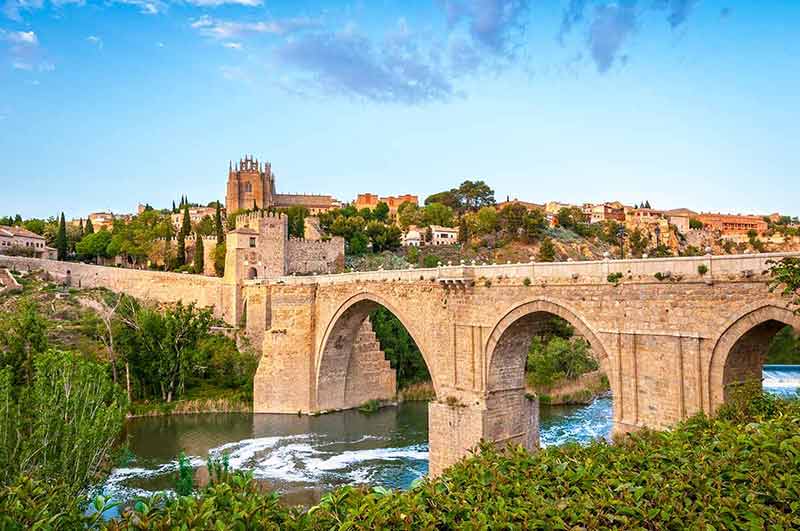 cities in spain by population the bridge Stone bridge across calm river. Blue sky reflected in crystal clear water. Big fort and country houses in background.