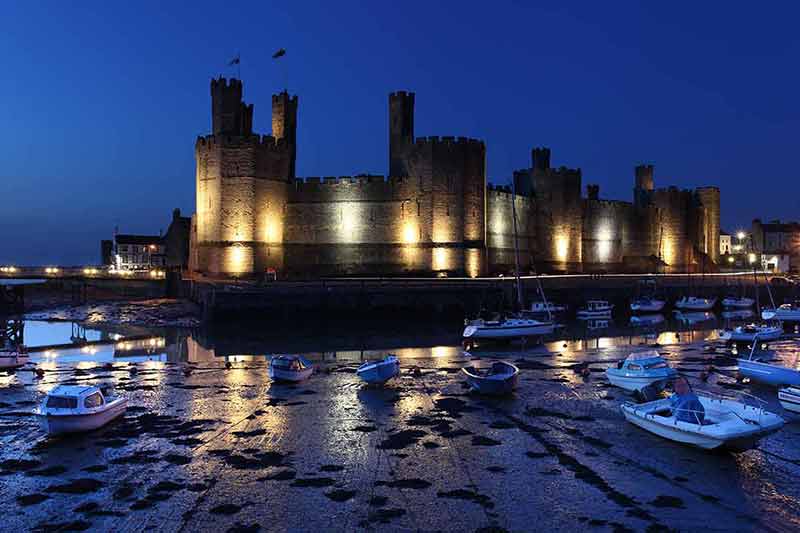 famous castles in wales Caernarfon castle lit up at night with boats in the foreground