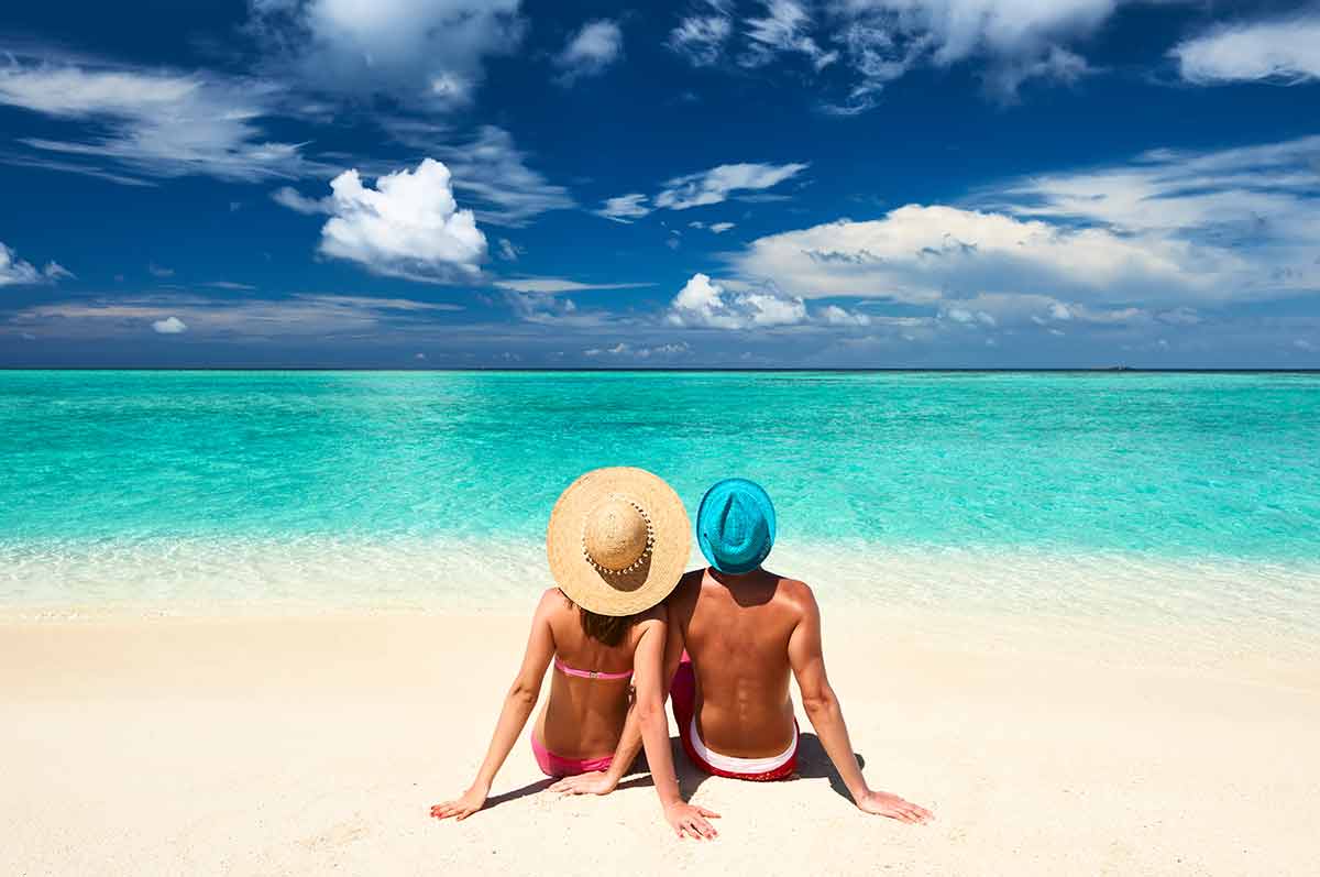 glowing beaches maldives man and woman sitting on the sand facing the ocean wearing hats