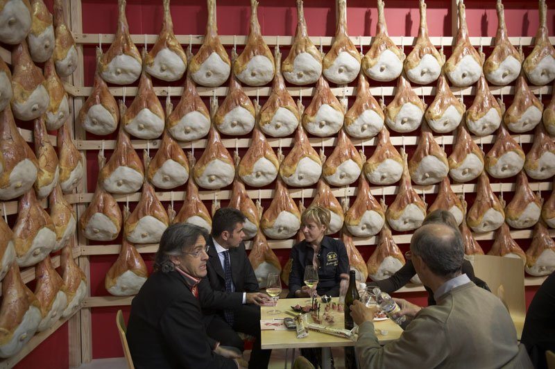 Visitors enjoying food and wine next to the leg ham wall at Salone del Gusto in Turin, Italy