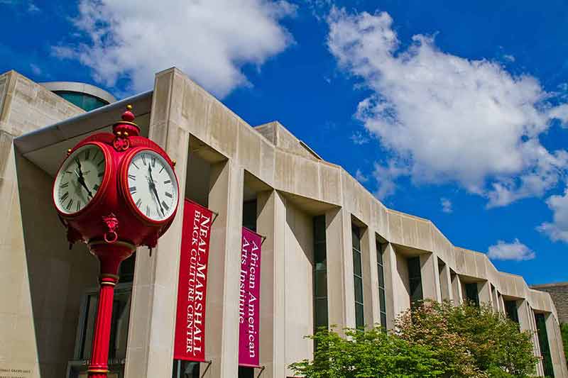 Image of Red antique clock in front of a white stone building with unique architecture Neal Marshall Auditorium at Indiana University in Bloomington.