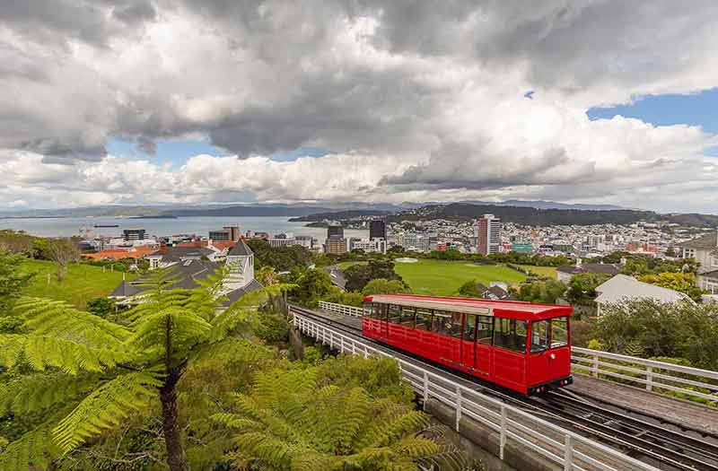 Beautiful blue sky covered with clouds. Red cable car moving up the mountain in the foreground and panorama of Wellington in the background.