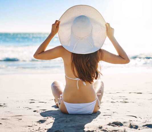 woman wearing a white hat and bikini sitting cross-legged on the beach