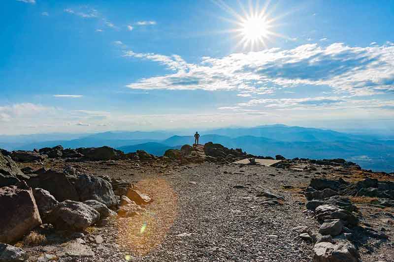 national parks in maine and new hampshire View from top Mount Washington across mountainous landscape to horizon under sun burst.