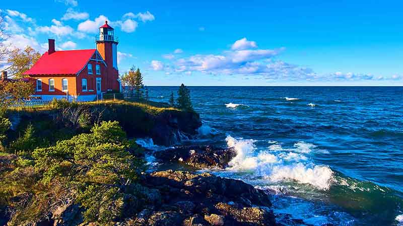 national parks near michigan red lighthouse with crashing lake waves and green trees with a gorgeous blue sky and white and purple clouds