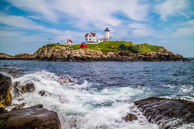 cape neddick nubble lighthouse across the water with waves crashing on the rocks in the foreground