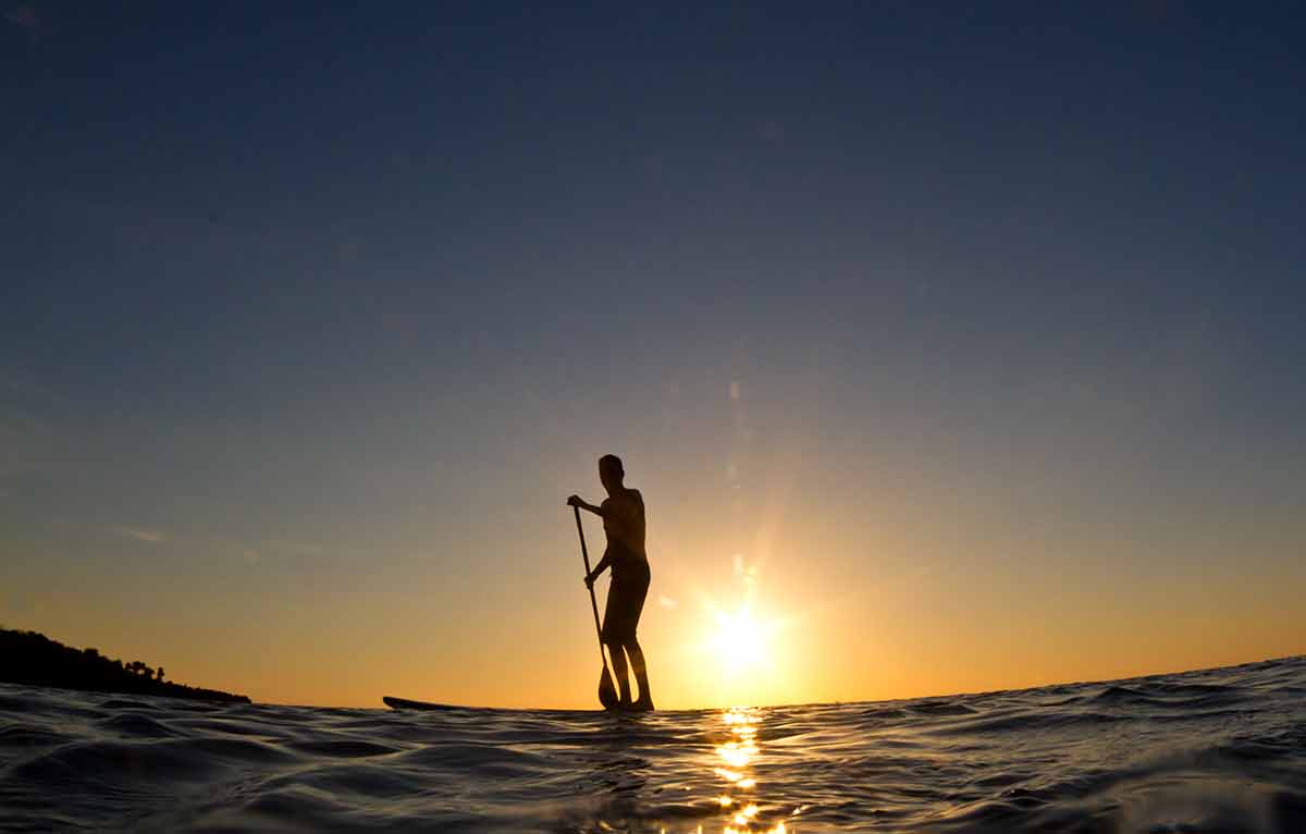 roatan bay islands honduras beaches A young man paddles his surfboard in to shore at sunset.