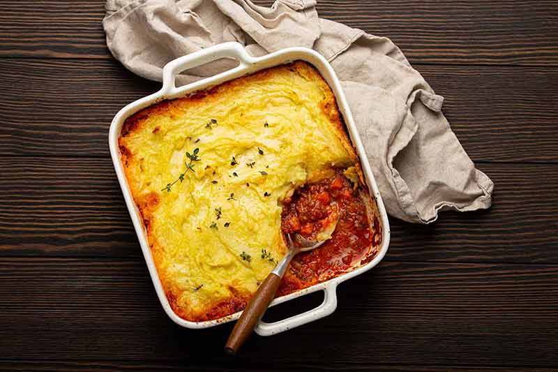 Shepherd's pie casserole with minced meat and mashed potatoes in ceramic baking dish on white rustic table with spoon.