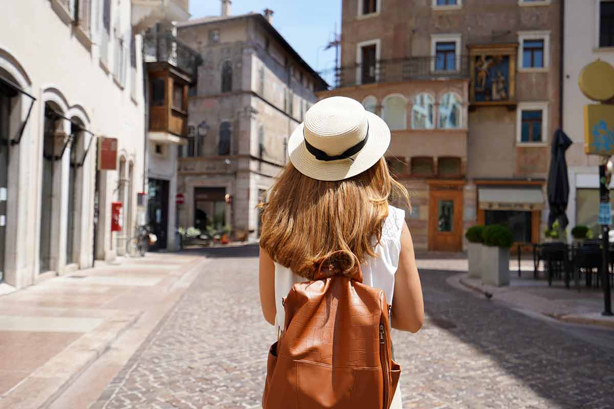young woman wearing a straw hat and tan backpack walking through a European town