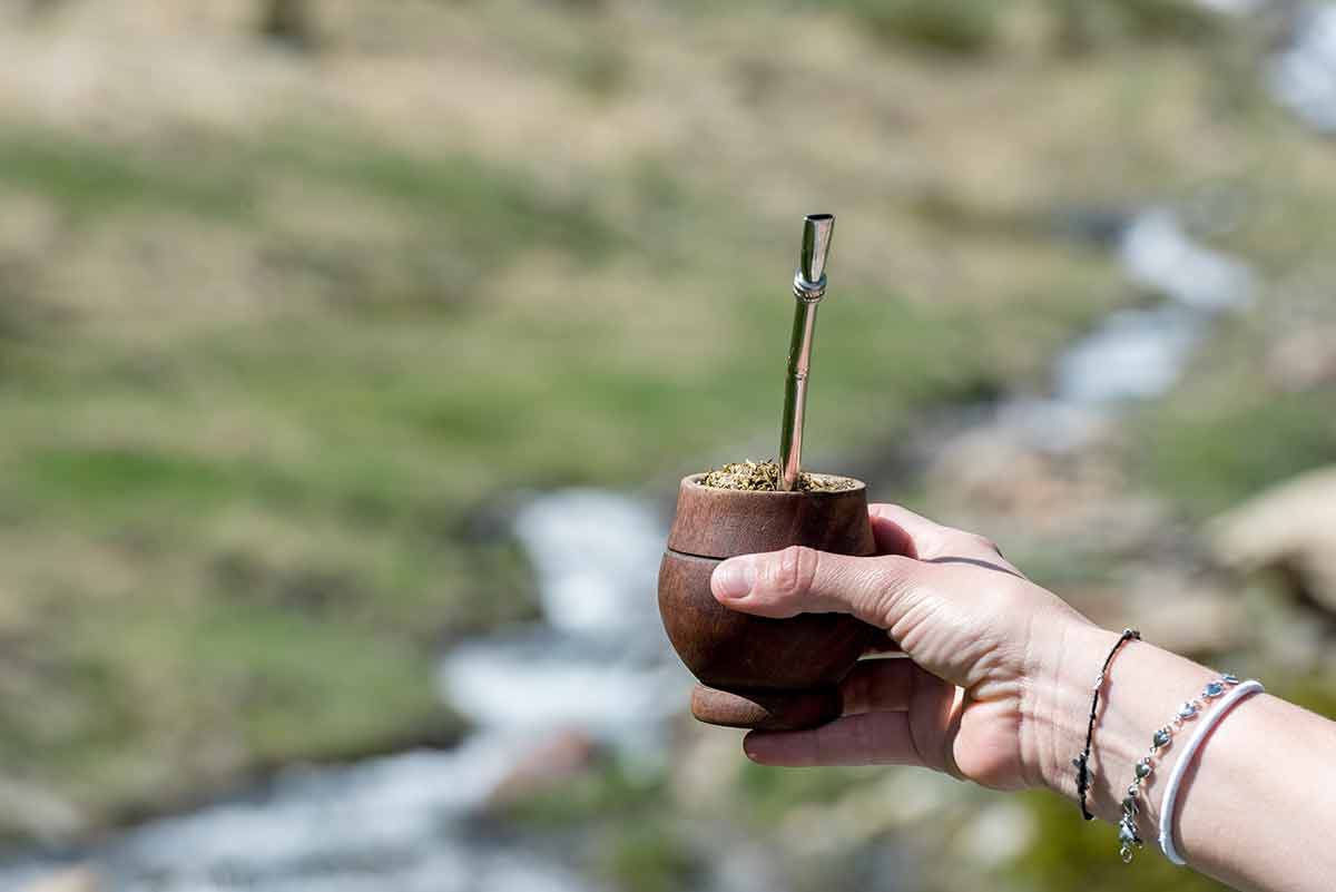 a hand holding a wooden cup with mate tea and a straw