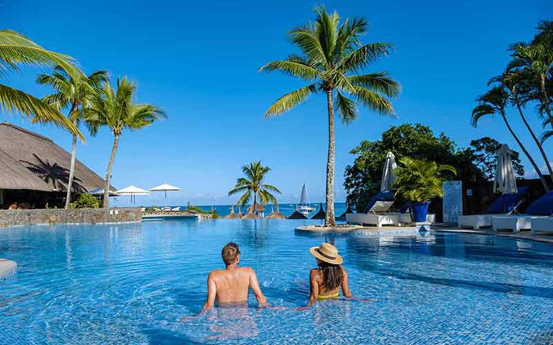 Beautiful tropical beachfront hotel resort with a swimming pool, sun-loungers, and palm trees, a paradise destination for vacations. Couple men and woman, mid age on a luxury vacation.