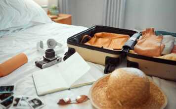 Travel, luggage and bedroom with a suitcase, hat and camera on a bed in a hotel during holiday or vacation. Hospitality, tourist and resort with luggage in a room for traveling or sightseeing abroad.
